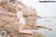 A woman in a white shirt and blue panties sitting on a rock by the water.